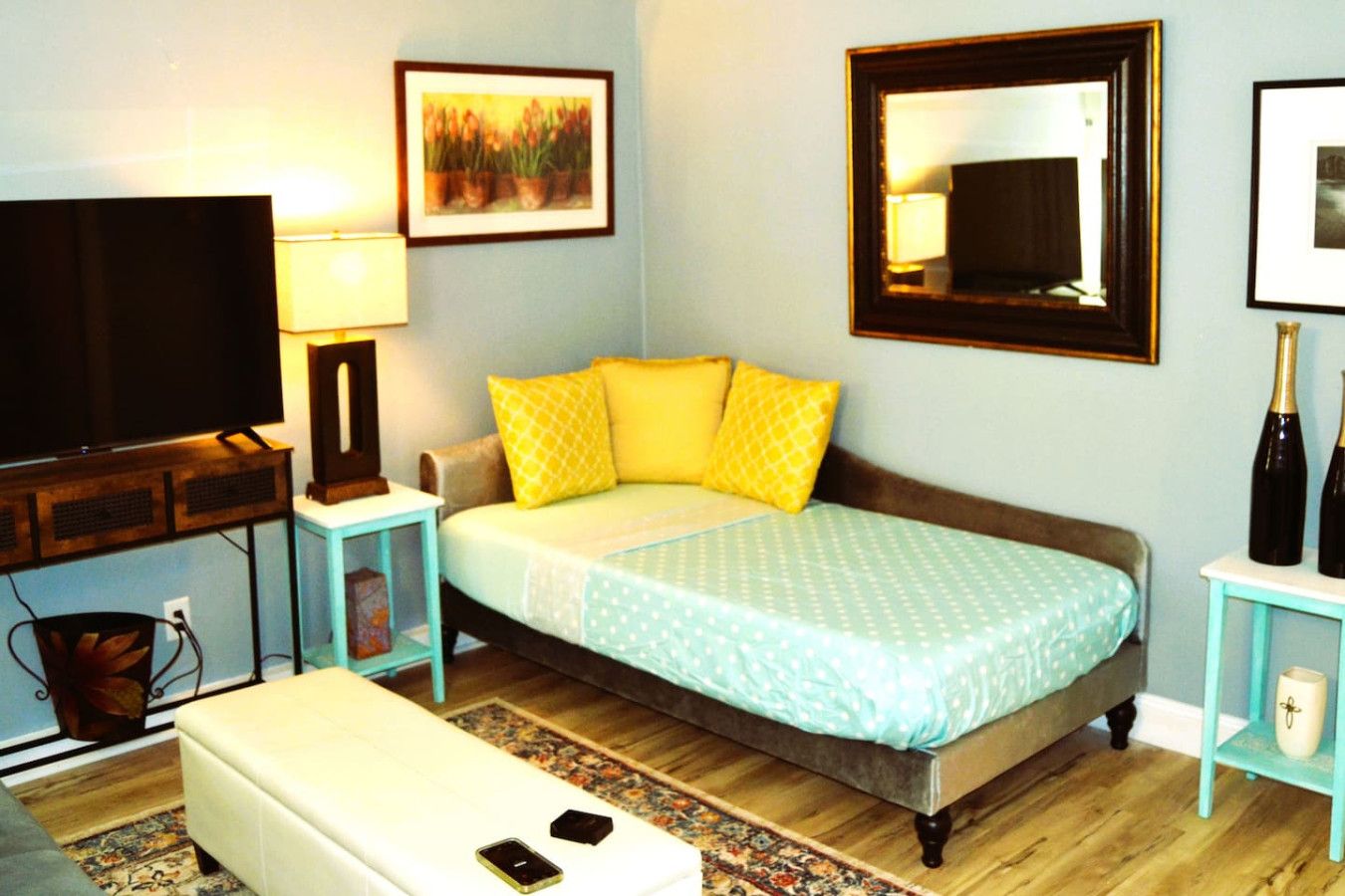 Un dormitorio con una cama, televisión, lámpara, marco de fotos y mesita de noche con botellas.
