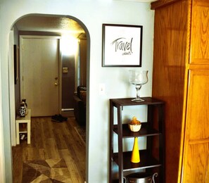 Um corredor com piso de madeira, uma porta, uma placa emoldurada na parede e uma pequena mesa com um copo e um vaso amarelo.