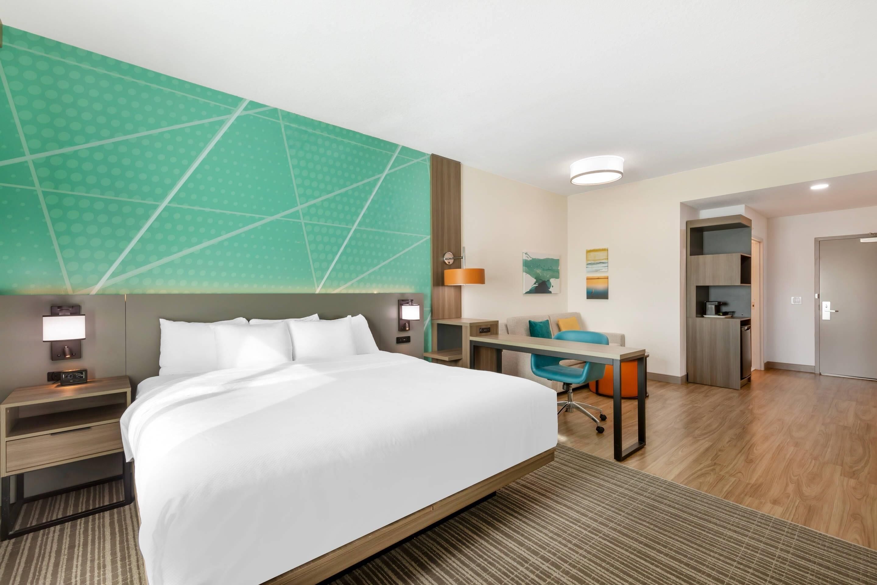 A hotel room with a large bed, a desk, a chair, a small kitchenette, and a bathroom.