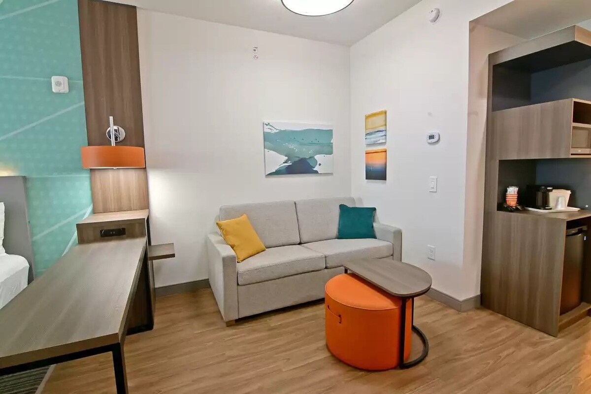 A modern room with a sofa, ottoman, and a small table. There is a kitchenette area with a microwave and a coffee maker.