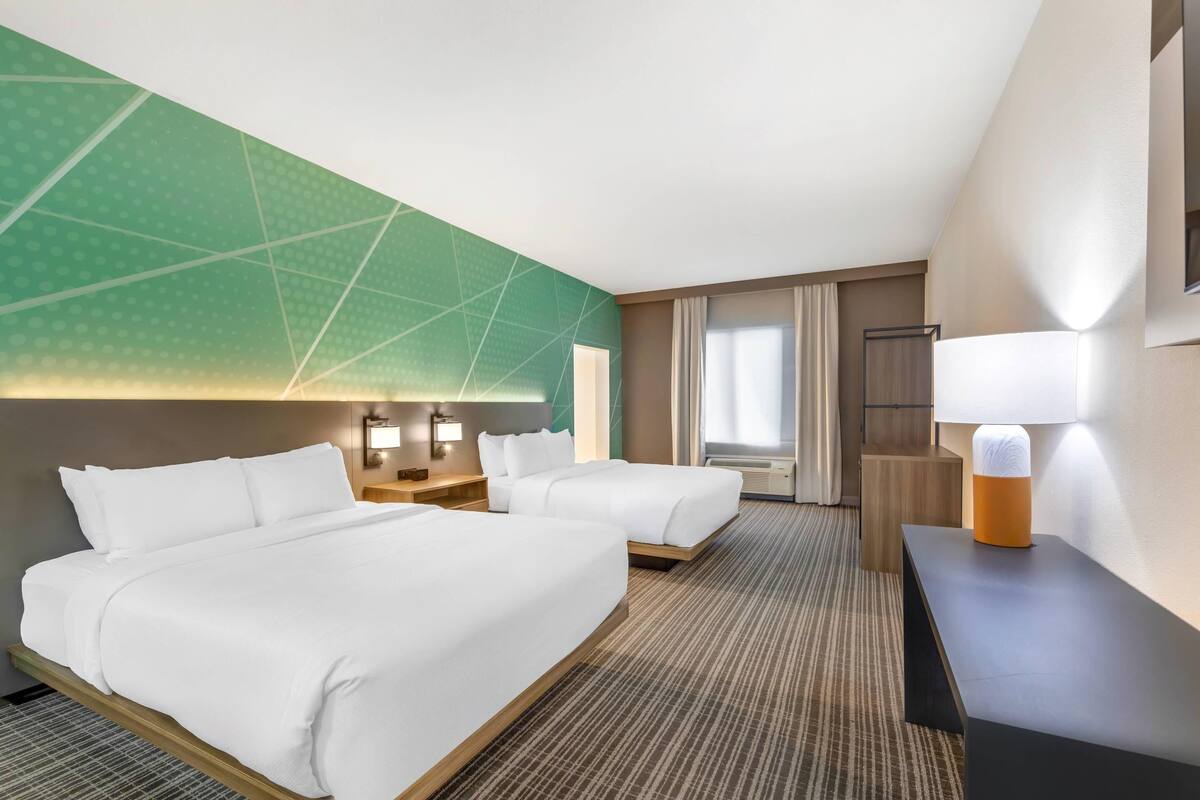 A hotel room with two beds, a desk, a lamp, and a large window with curtains.