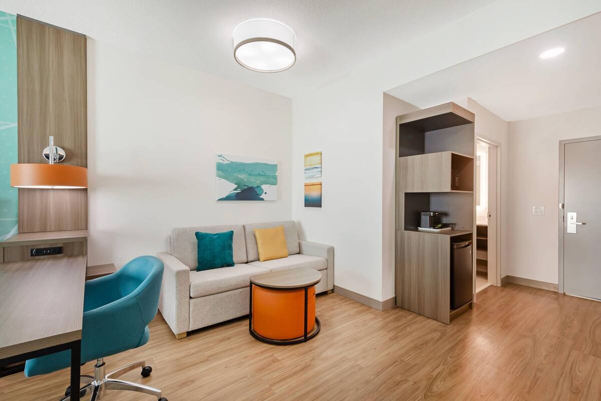 A modern living room with a sofa, a round orange table, a blue chair, and a kitchen area with built-in shelves.