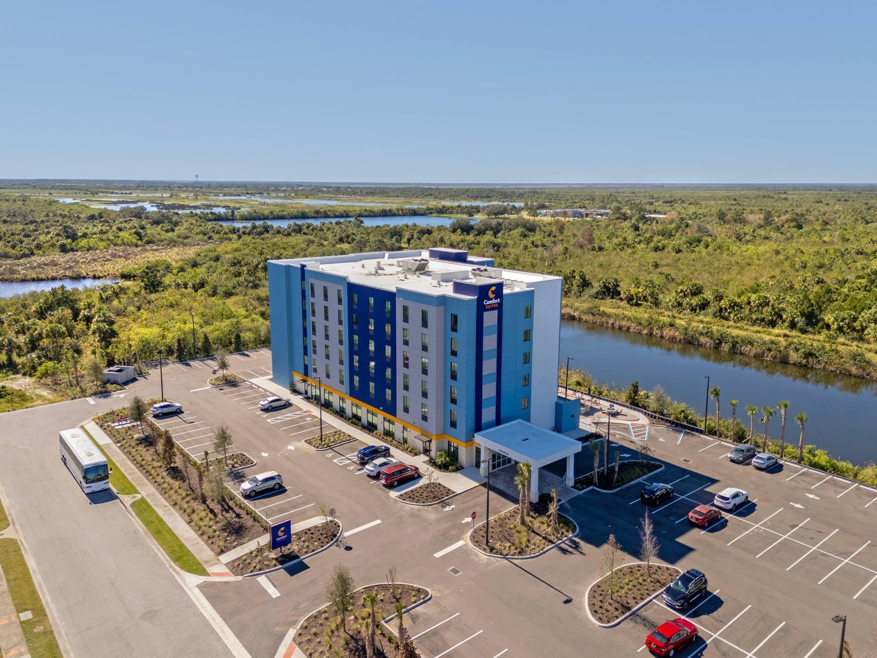A hotel with a parking lot, a building with multiple floors, and a body of water in the background.