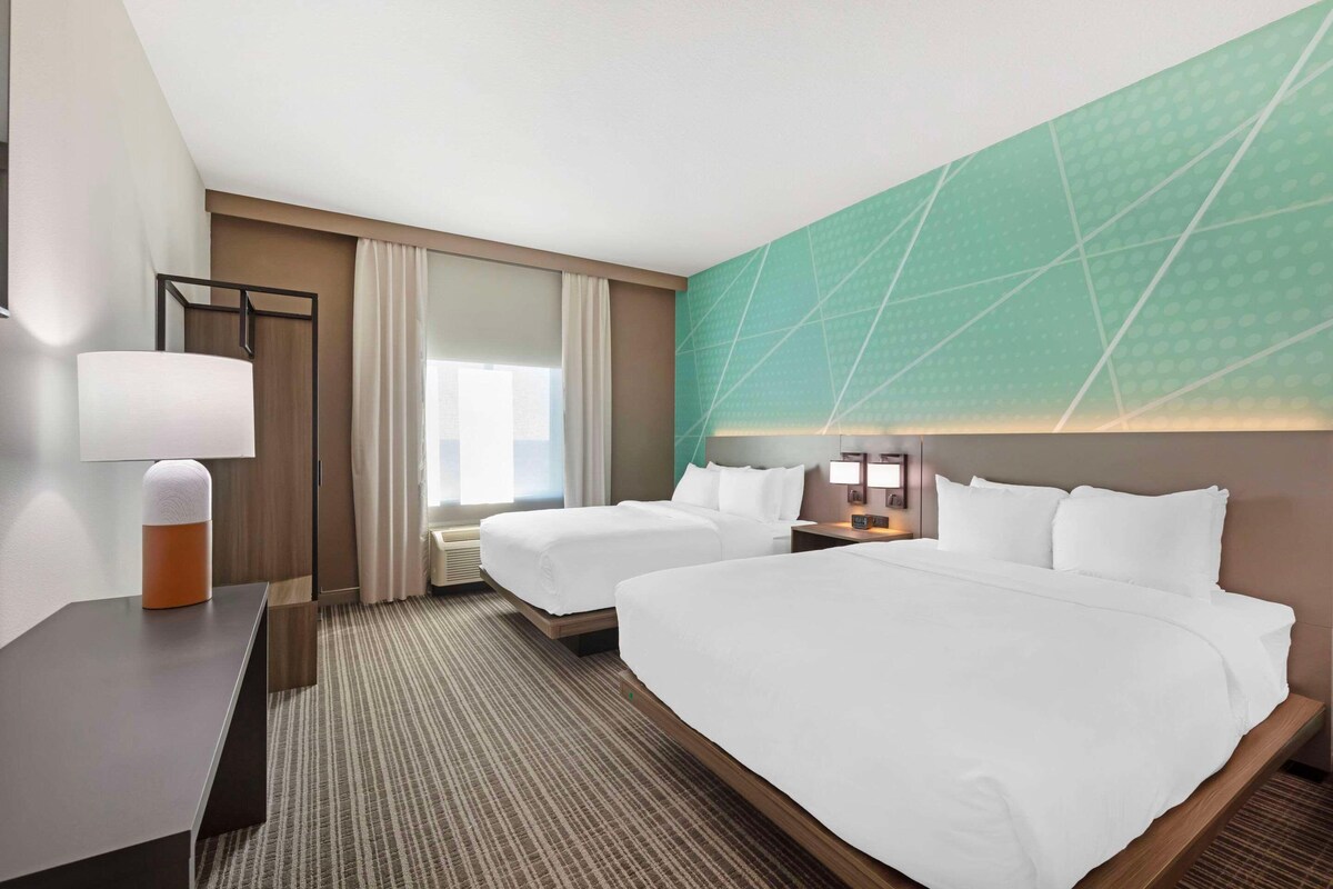 A hotel room with two beds, a desk, a lamp, and a large window with curtains.