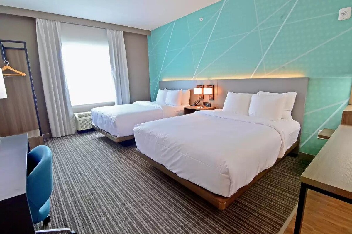 Ein Hotelzimmer mit zwei Betten, einem Schreibtisch, einem Stuhl, einer Schreibtischlampe und einem Fenster mit Vorhängen.
