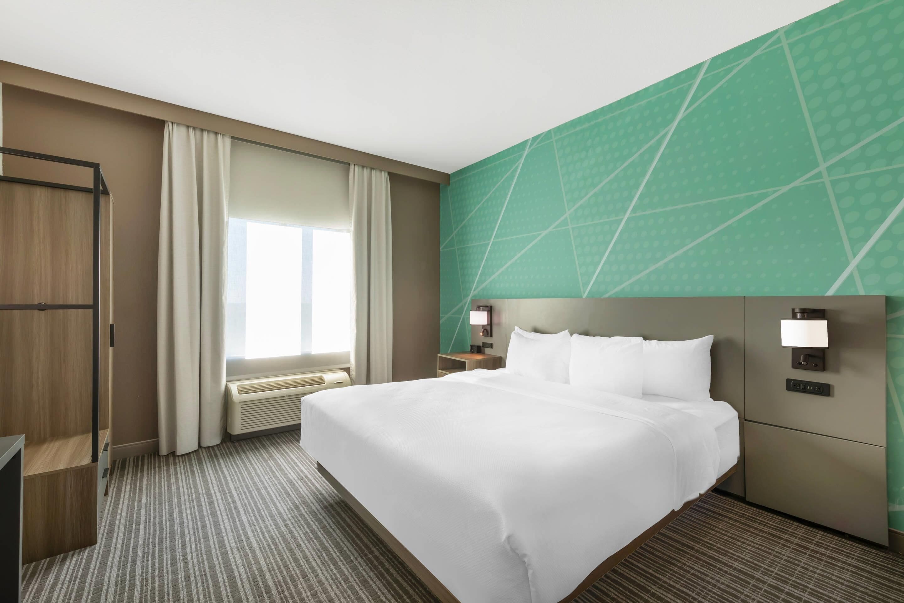 A hotel room with a large bed, a nightstand, a desk, a wardrobe, and a window with curtains.