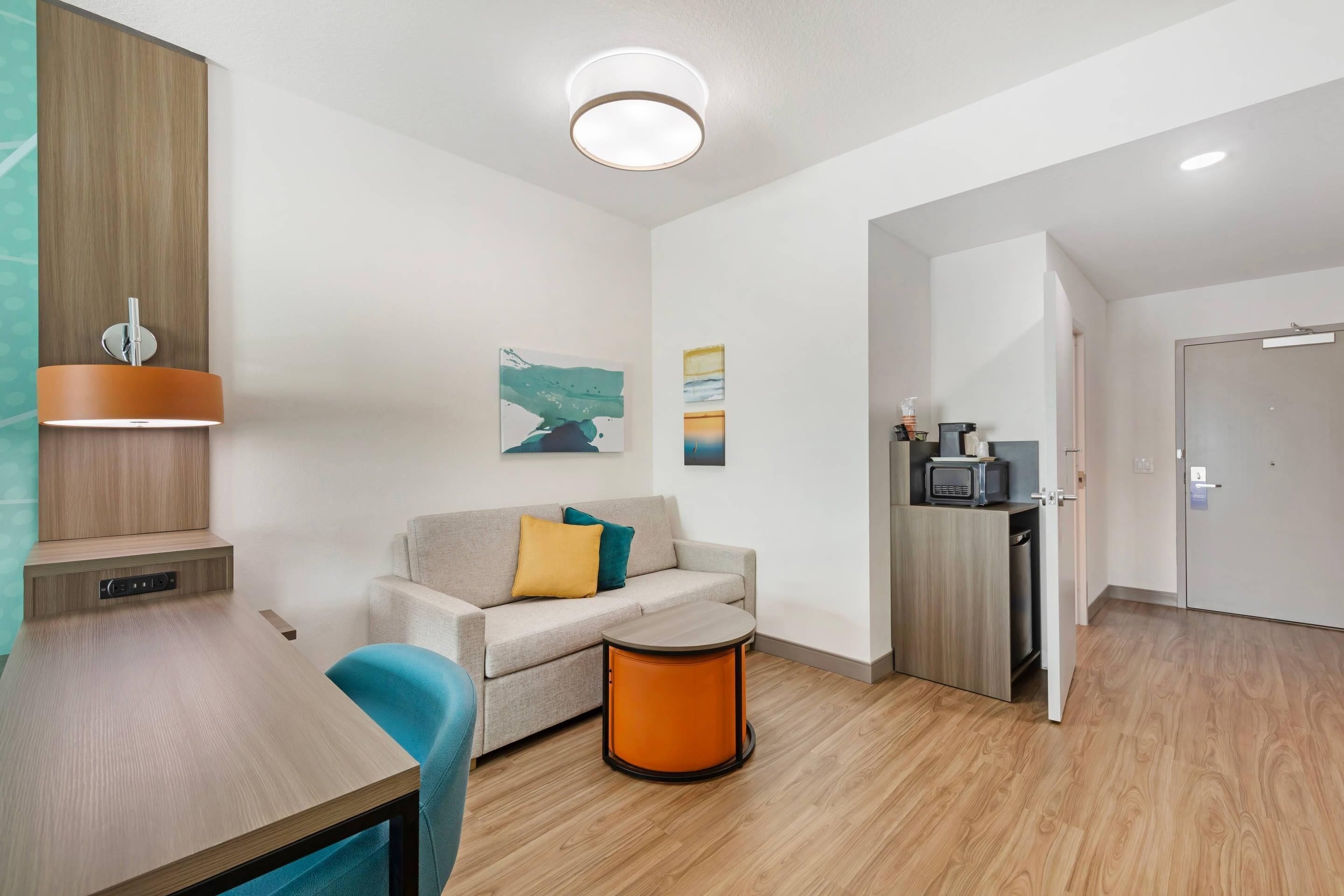 Habitación de hotel moderna con piso de madera, un sofá con cojines, una mesita redonda pequeña, una kitchenette con microondas y una puerta que conduce a otra habitación.