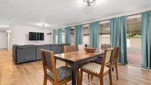 A modern living room with a dining table, wooden chairs, a sofa, and a TV mounted on the wall. - Eastside Family Summer Retreat | Yard & Games (Tucson)