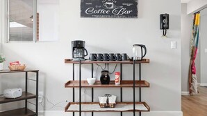 A coffee bar setup with a coffee maker, cups, and a coffee pot on a shelf, a bowl of snacks, and a coffee jar on the floor. - Eastside Family Summer Retreat | Yard & Games (Tucson)
