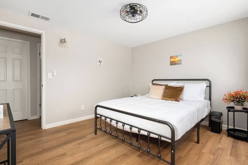 Una camera da letto con una struttura del letto in metallo, biancheria da letto bianca, un piccolo comodino con un vaso di fiori e una ventola a soffitto.