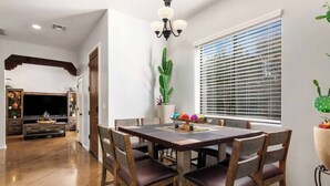 A dining area with a wooden table and chairs, a cactus plant, and a window with blinds. - Cozy Summer Oasis | Near UofA & Shows (Tucson)