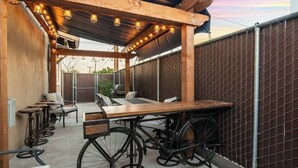 An outdoor patio with a wooden table and chairs, a bicycle, and a pergola with lights. - Cozy Summer Oasis | Near UofA & Shows (Tucson)