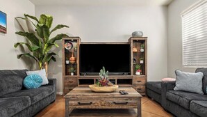 A living room with a large flat-screen TV, a coffee table with decorative items, two sofas with cushions, and a large plant. - Cozy Summer Oasis | Near UofA & Shows (Tucson)