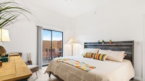 Un dormitorio con una cama grande, un escritorio con lámpara y vistas al exterior a través de un ventanal corredera.