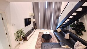 A modern living room with a black leather sofa, a coffee table, and a TV mounted on the wall. There is a staircase on the right side and a potted plant on the left. - The Gem of Embassy Gardens  (Accra)
