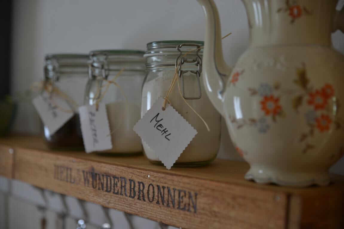 A shelf with glass jars and a ceramic pitcher, each labeled with handwritten tags.