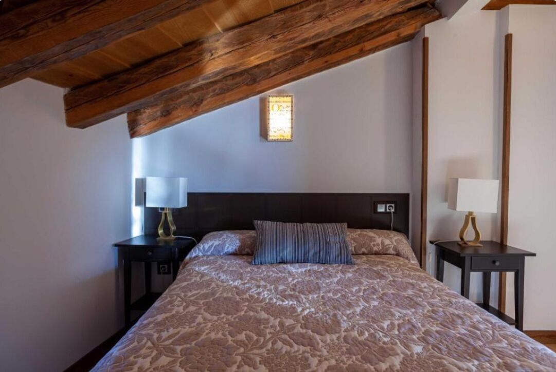 A bedroom with a large bed, wooden beams, two bedside tables with lamps, and a small wall-mounted light.