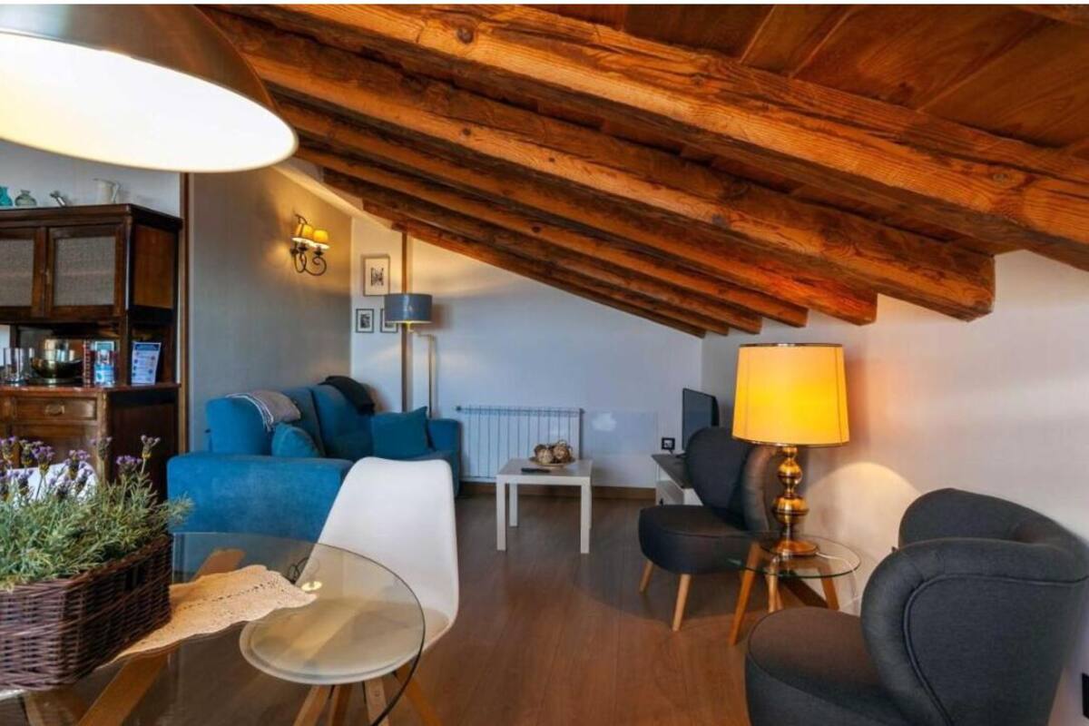 A cozy loft-style living room with a wooden ceiling, a blue sofa, a white armchair, a small table, and a lamp.