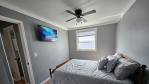 Une chambre à coucher avec un lit, un ventilateur de plafond, un téléviseur fixé au mur et une fenêtre.