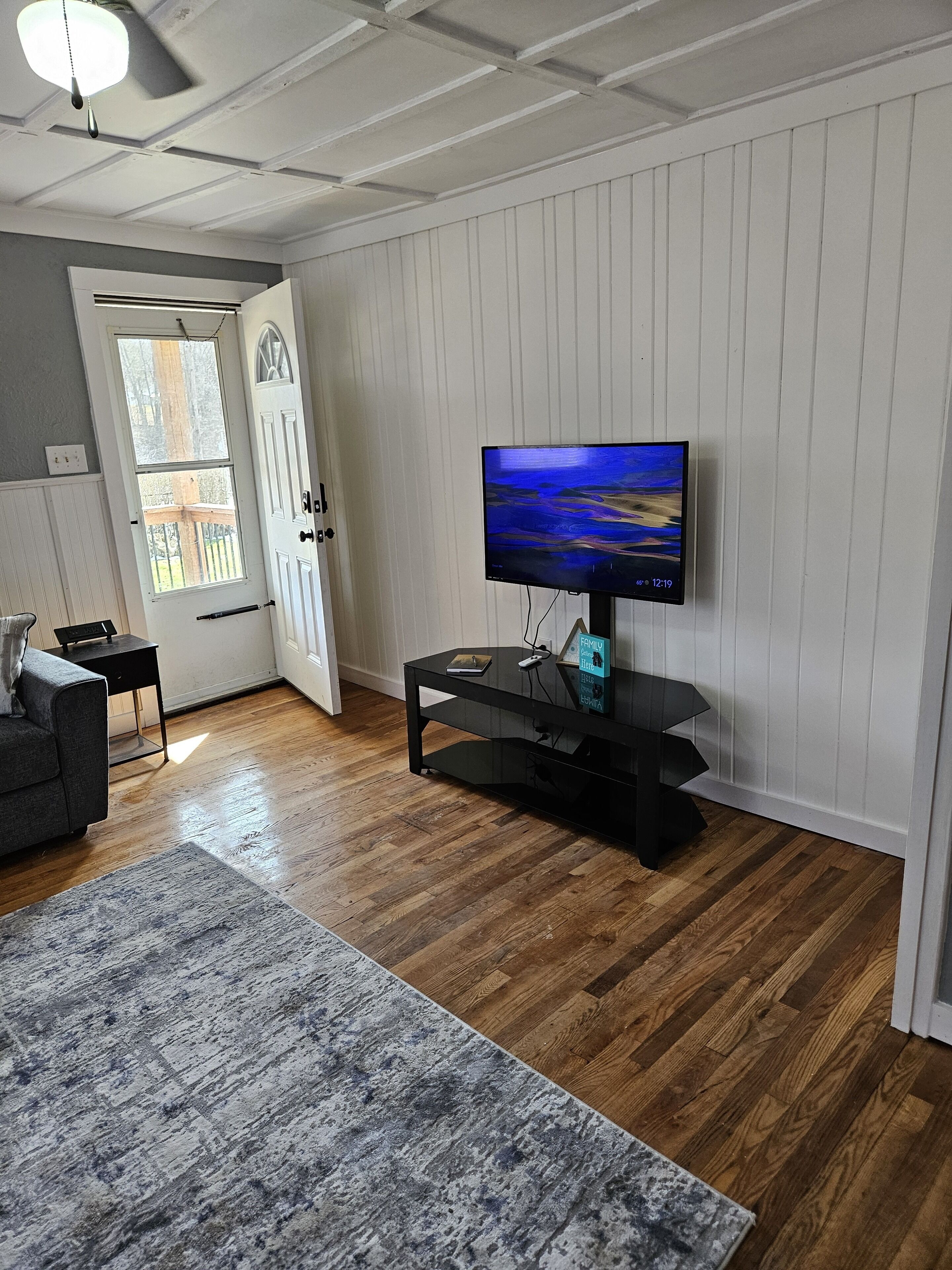 A living room with a flat-screen TV on a black stand, a grey sofa, a small side table, and a wooden floor.