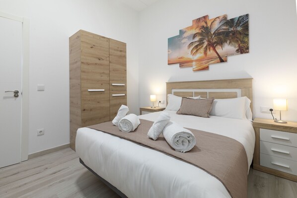 A hotel room with a bed, a wooden wardrobe, a nightstand with a lamp, and a wall with a tropical sunset painting.