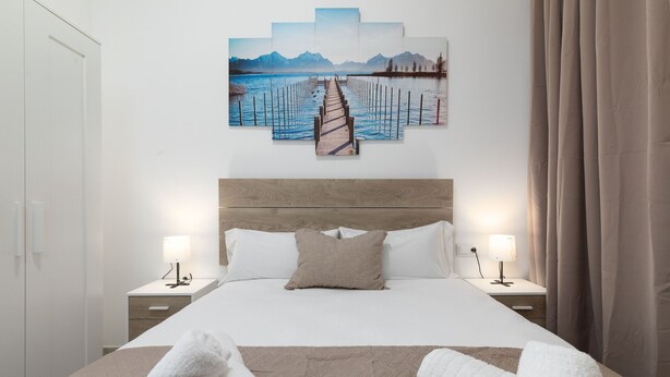 A neatly made bed with white linens, two bedside tables with lamps, and a large wall art piece depicting a pier leading to mountains. - Malvarrosa Las Arenas Access. WiFi + Air Conditioning + 5 Pax (Valencia)