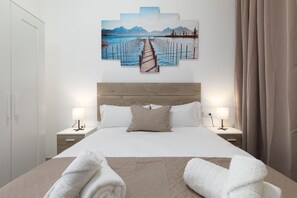 A neatly made bed with white linens, two bedside tables with lamps, and a large wall art piece depicting a pier leading to mountains.