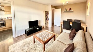Una sala moderna con sofá, mesa de centro y televisor. Se aprecia una cocina con alacenas y estufa. También se ve un comedor con sillas y mesa.