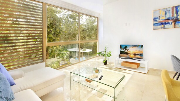 Un salon moderne avec une table basse en verre, un canapé blanc, un téléviseur posé sur un meuble blanc et de grandes fenêtres équipées de stores.