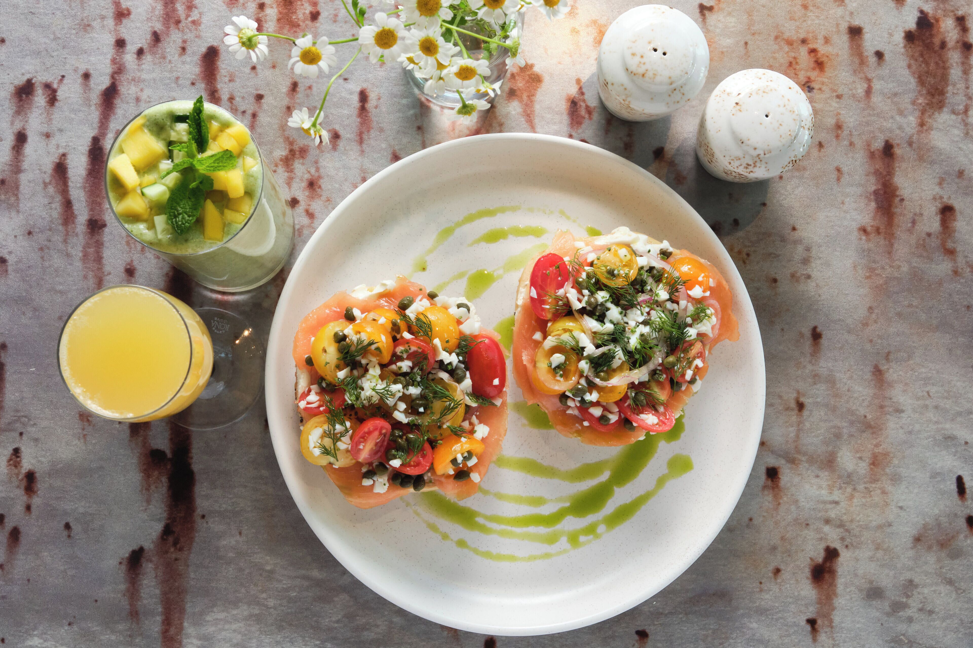 A plate with two open-faced sandwiches topped with various ingredients, a glass of orange juice, and a green drink with mint.