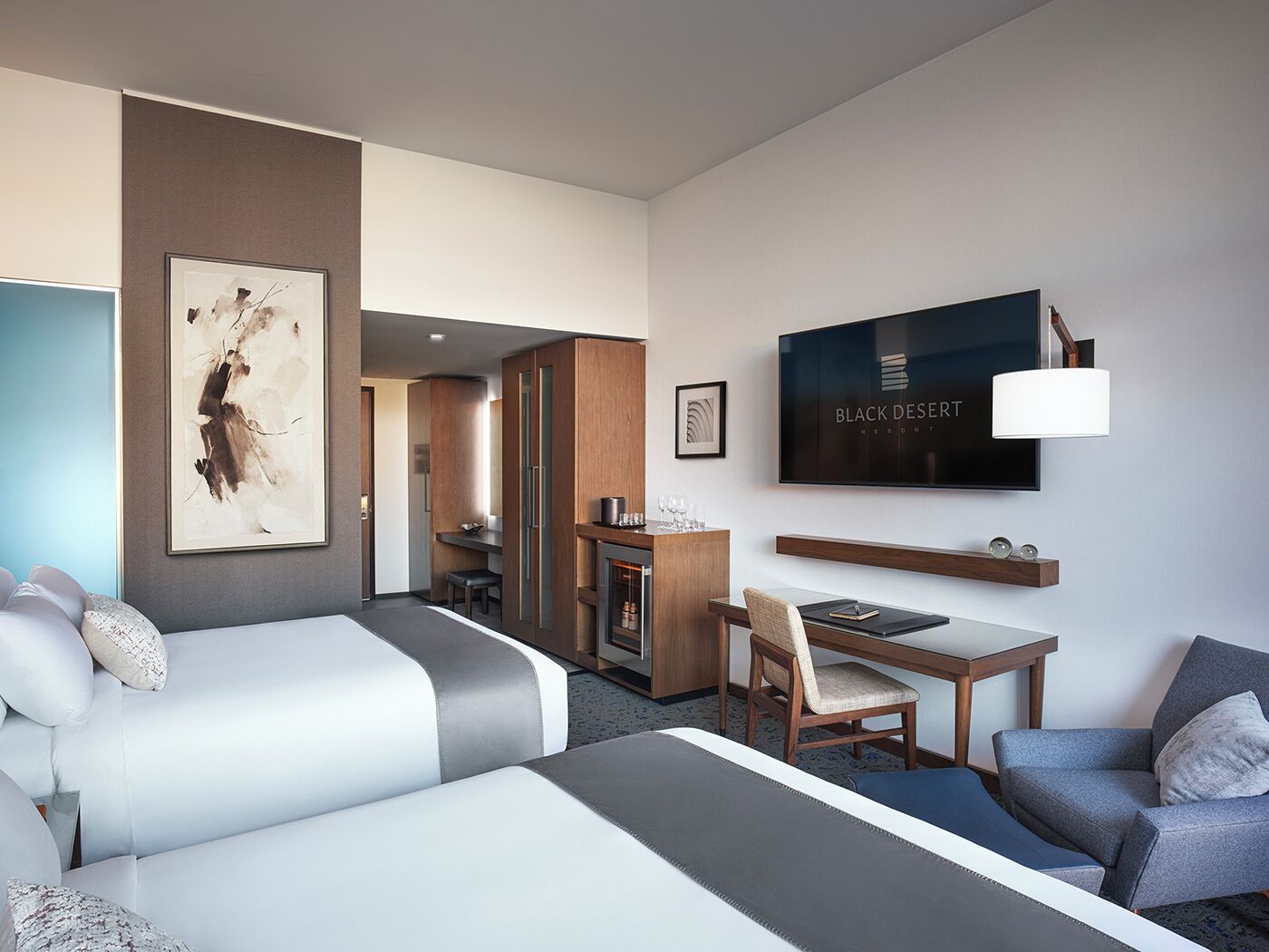 A hotel room with two beds, a desk, a chair, a TV, and a framed picture on the wall.