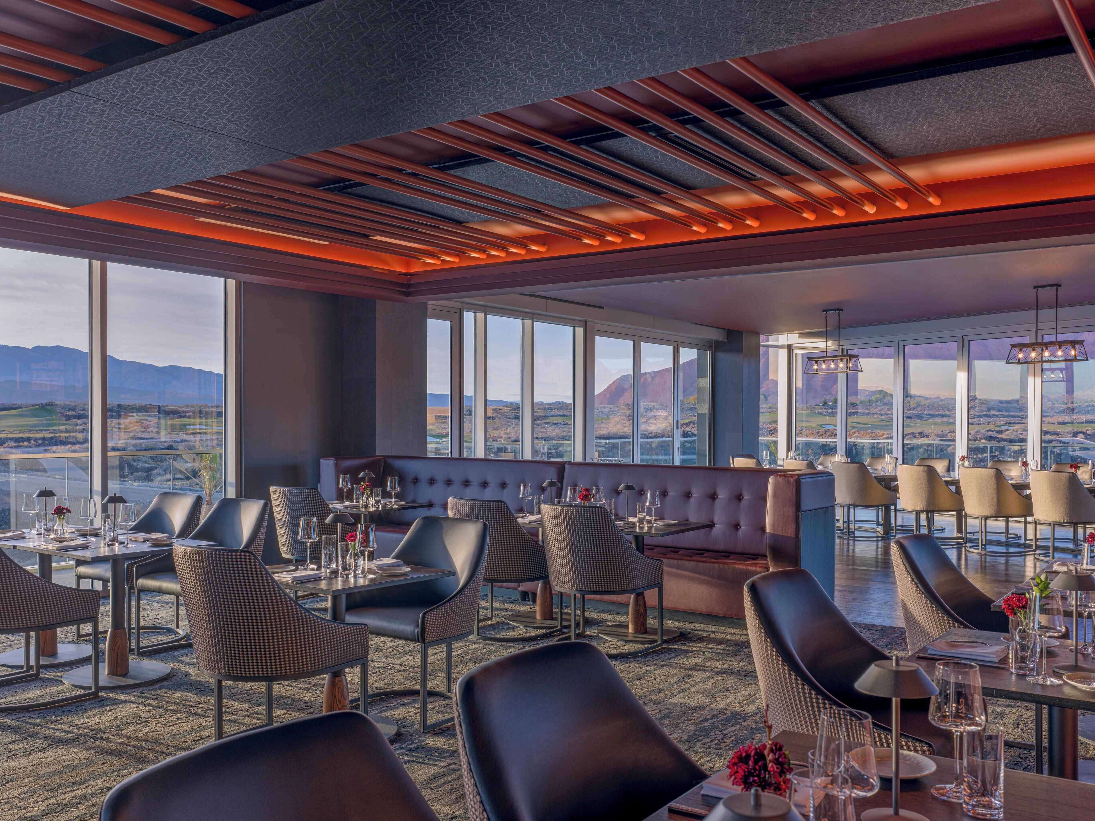 A modern restaurant with a panoramic mountain view, featuring a mix of leather and woven chairs, and a variety of table settings.