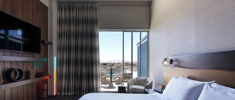 A modern hotel room with a large bed, a television, a view of the desert, and a balcony with seating.