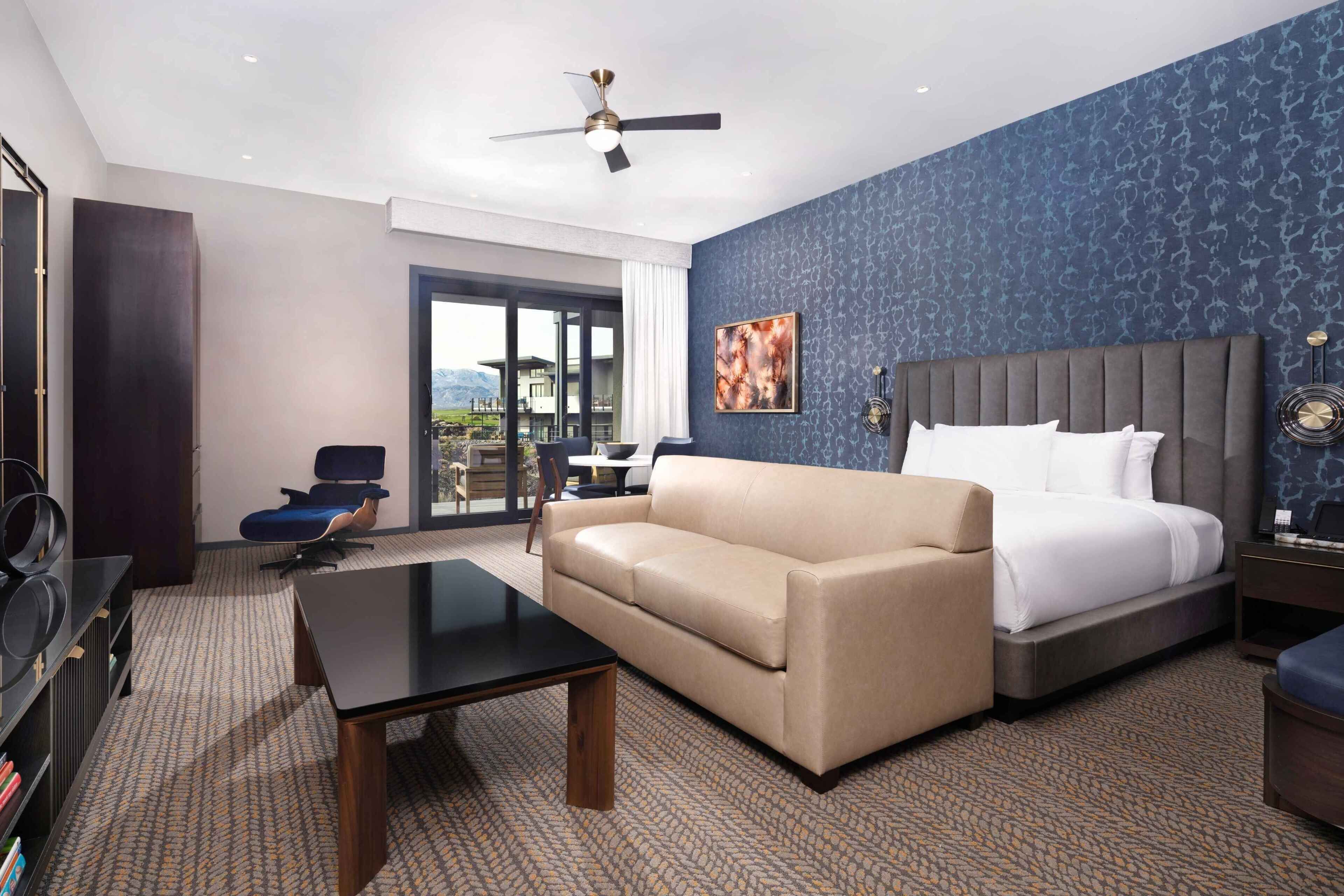 A hotel room with a bed, a sofa, a desk, a chair, a ceiling fan, and a view of the outdoors.