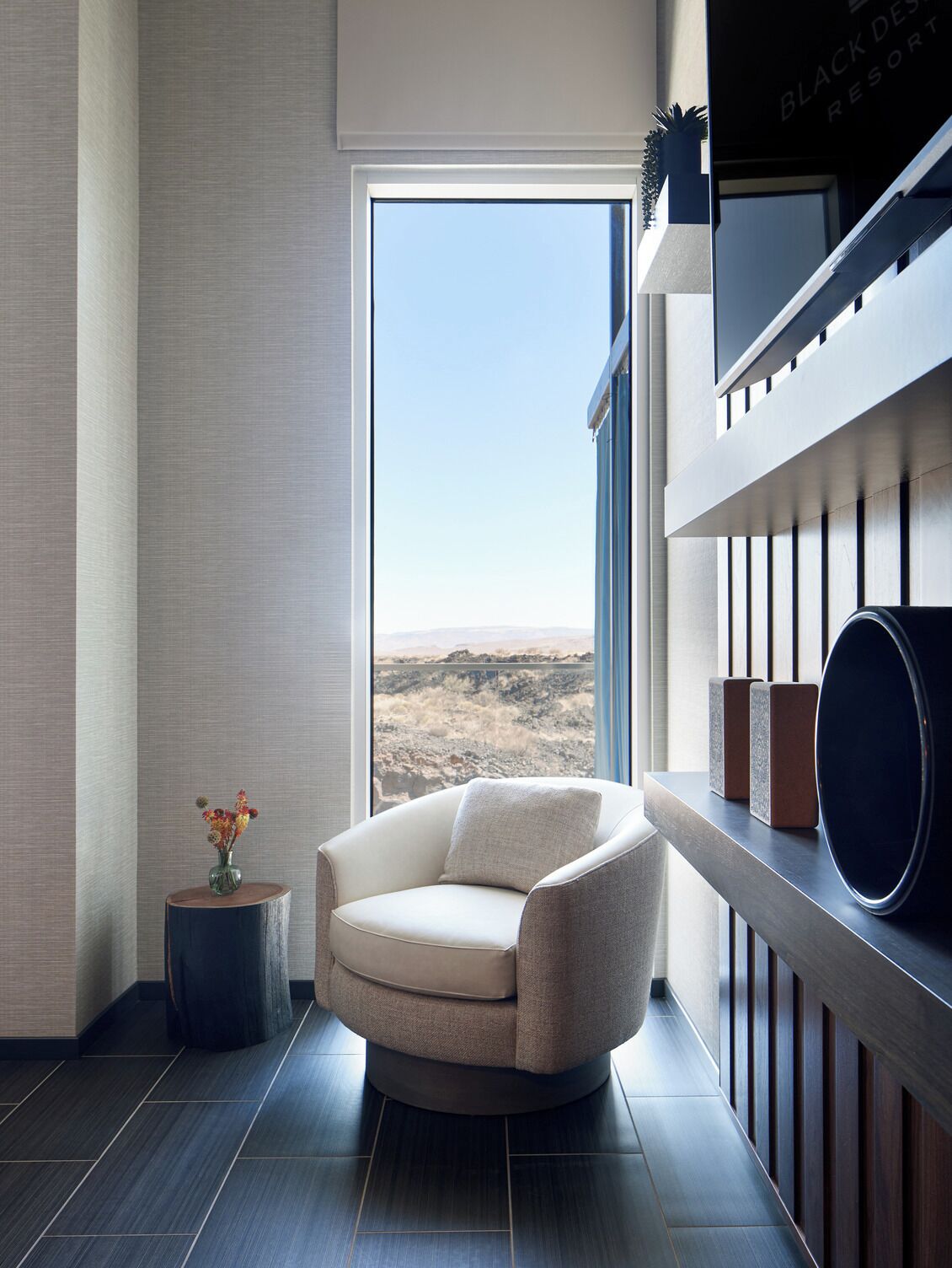Una habitación moderna con un ventanal grande, una silla redonda, una mesita auxiliar con un florero y un televisor montado.