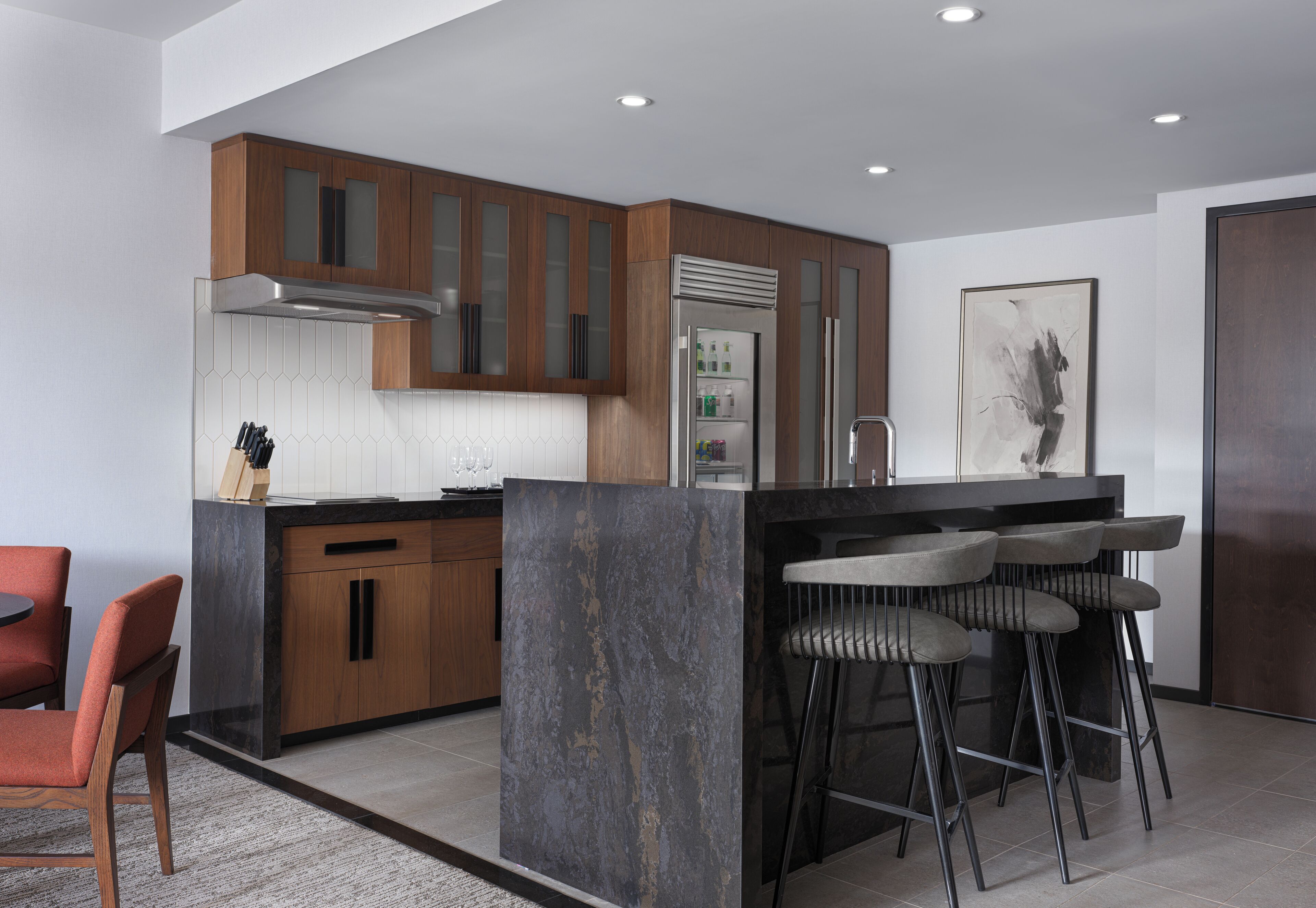 A modern kitchen with wooden cabinets, a stainless steel range hood, a refrigerator, and a bar area with stools.