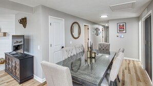 A dining area with a glass-top table, cushioned chairs, a dark wooden cabinet, and a wall-mounted mirror. - Remote Work Retreat: 3BR w/ Workspace Near KC! (Olathe)
