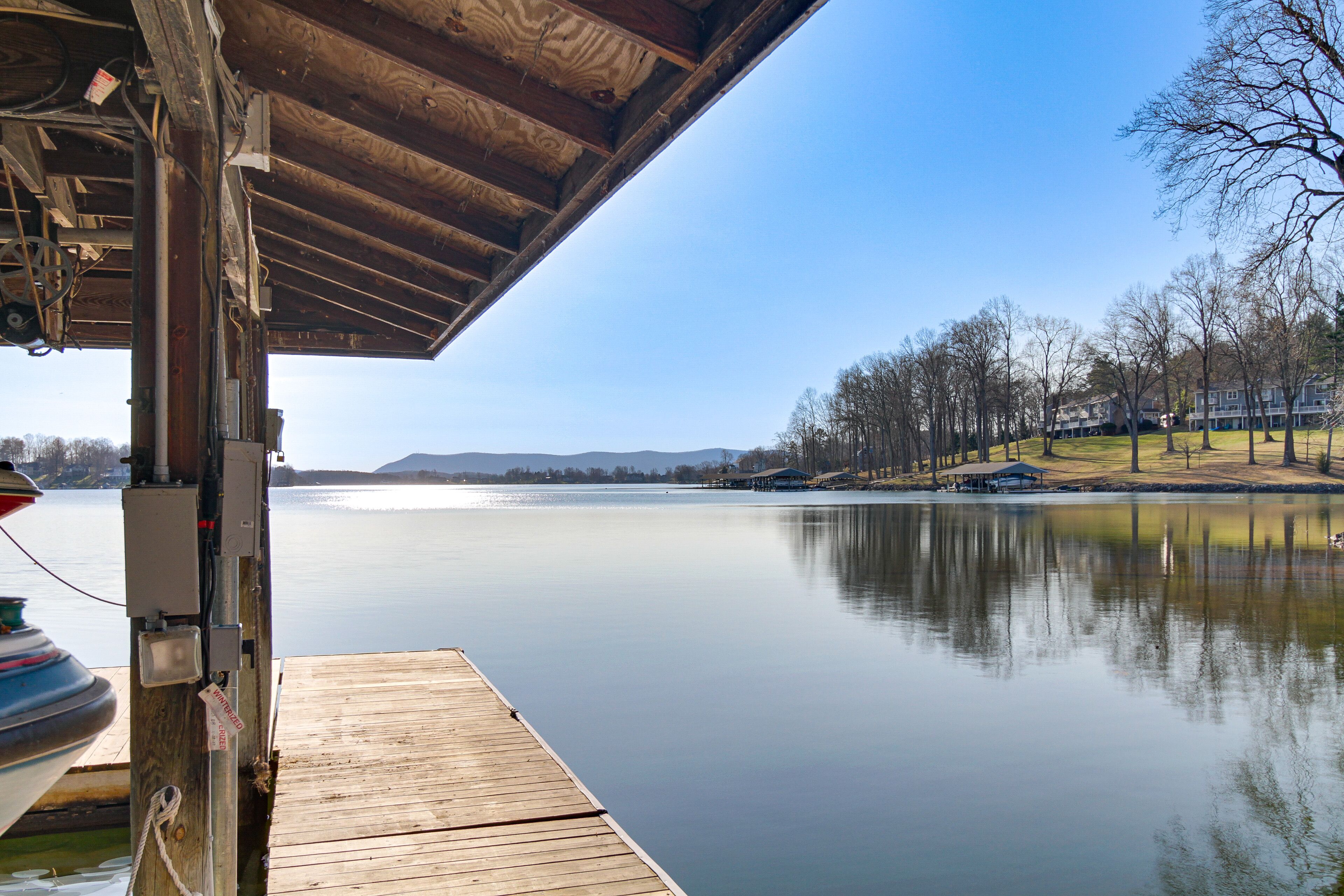 Un muelle de madera se adentra en un lago tranquilo, con un cielo despejado y edificios a lo lejos.