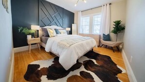 Un dormitorio moderno con una pared de acento oscura, un tapete de cuero de vaca, una cama con ropa blanca y un diseño geométrico en la pared.