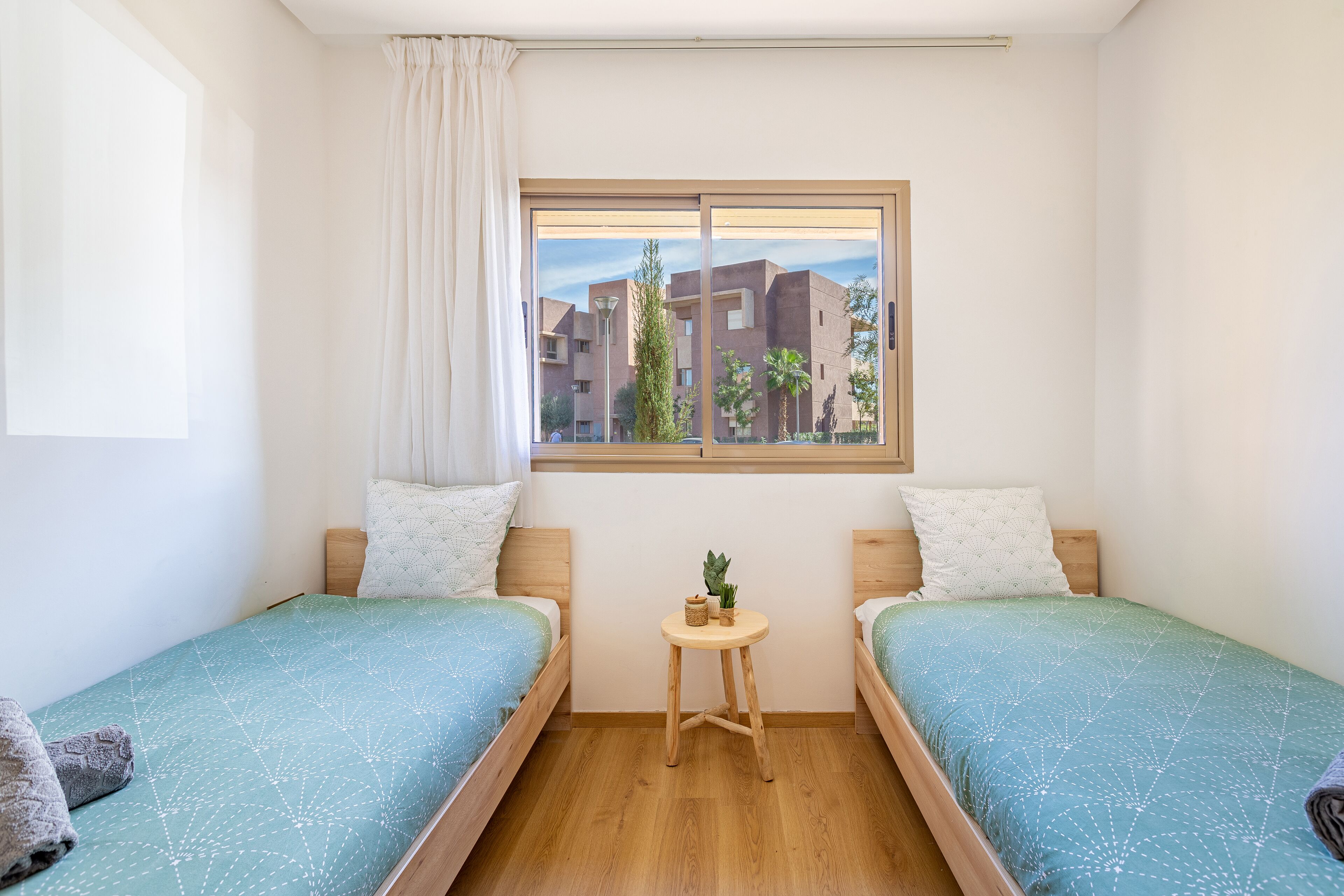 Une chambre avec deux lits simples, une petite table avec une plante et une fenêtre donnant sur des immeubles.