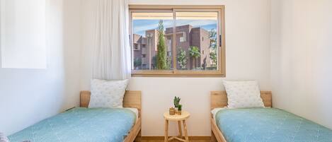 Une chambre avec deux lits simples, une petite table avec une plante et une fenêtre donnant sur des immeubles.