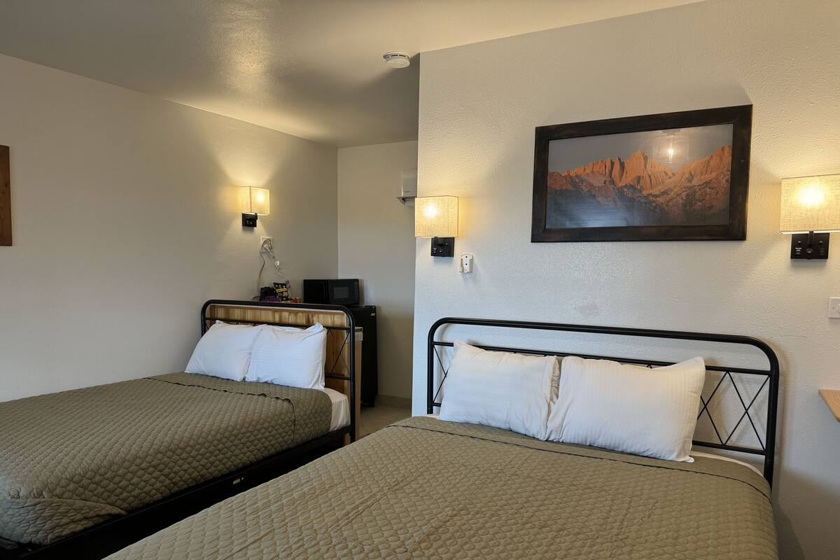 A hotel room with two beds, each with a quilted mattress and white pillows. There are wall-mounted lamps on either side of the beds, and a framed picture above the right bed.