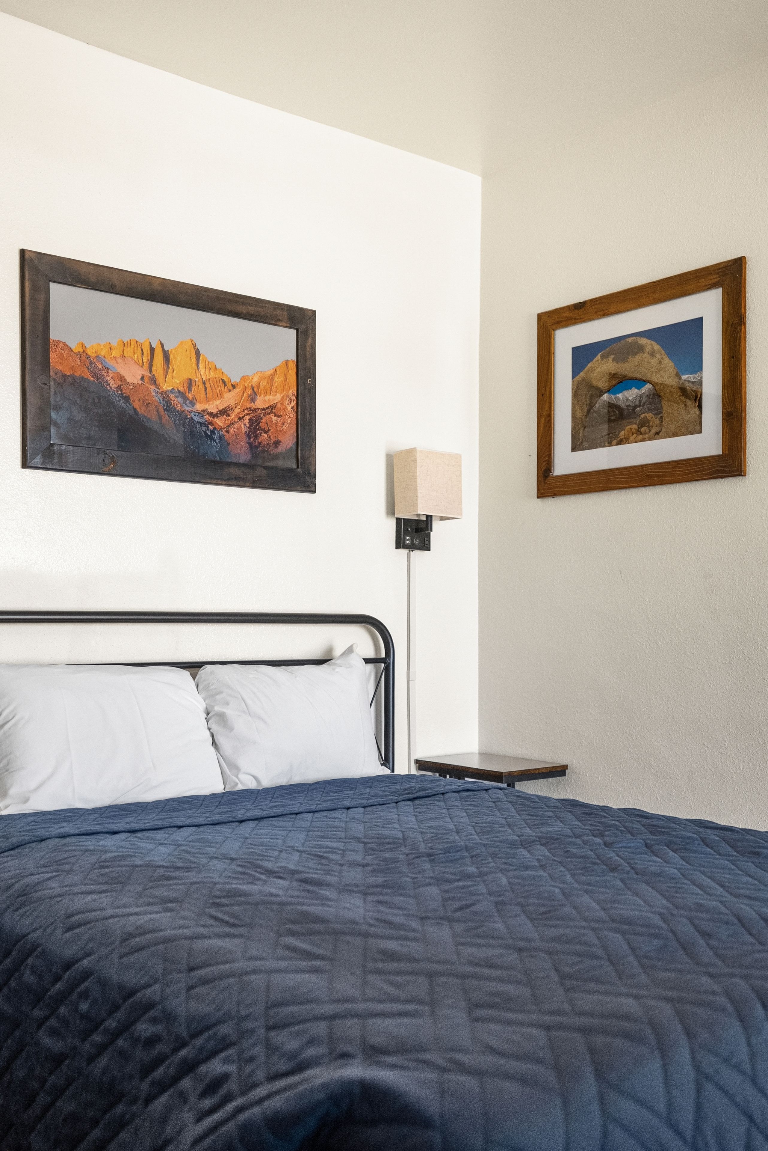 Una cama con colcha azul, dos almohadas blancas y un cabecero de madera. Hay dos cuadros en la pared, uno encima de la cama y otro a la derecha.