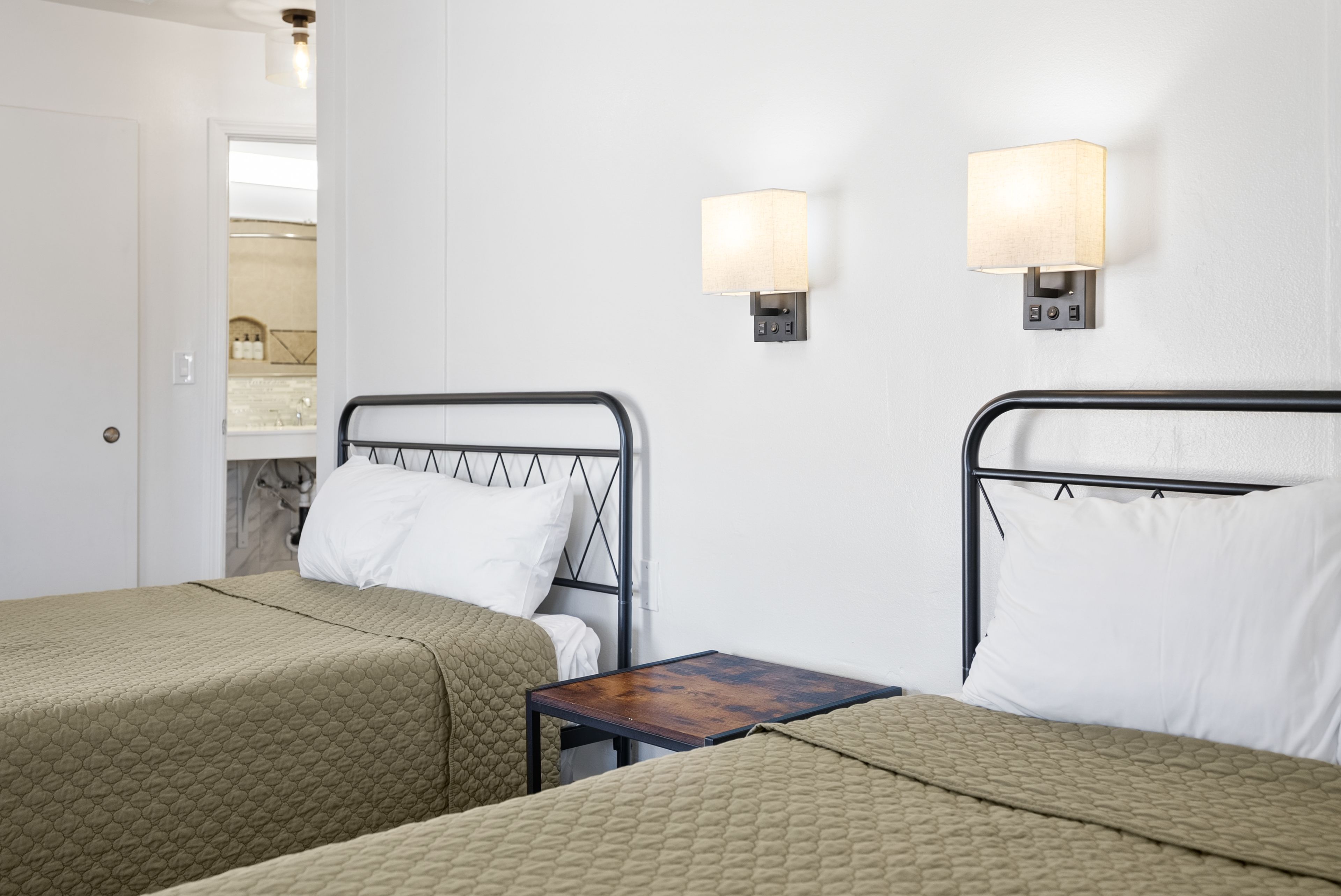 A hotel room with two single beds, each with a headboard and a bedside table. There are wall-mounted lamps on either side of the beds.