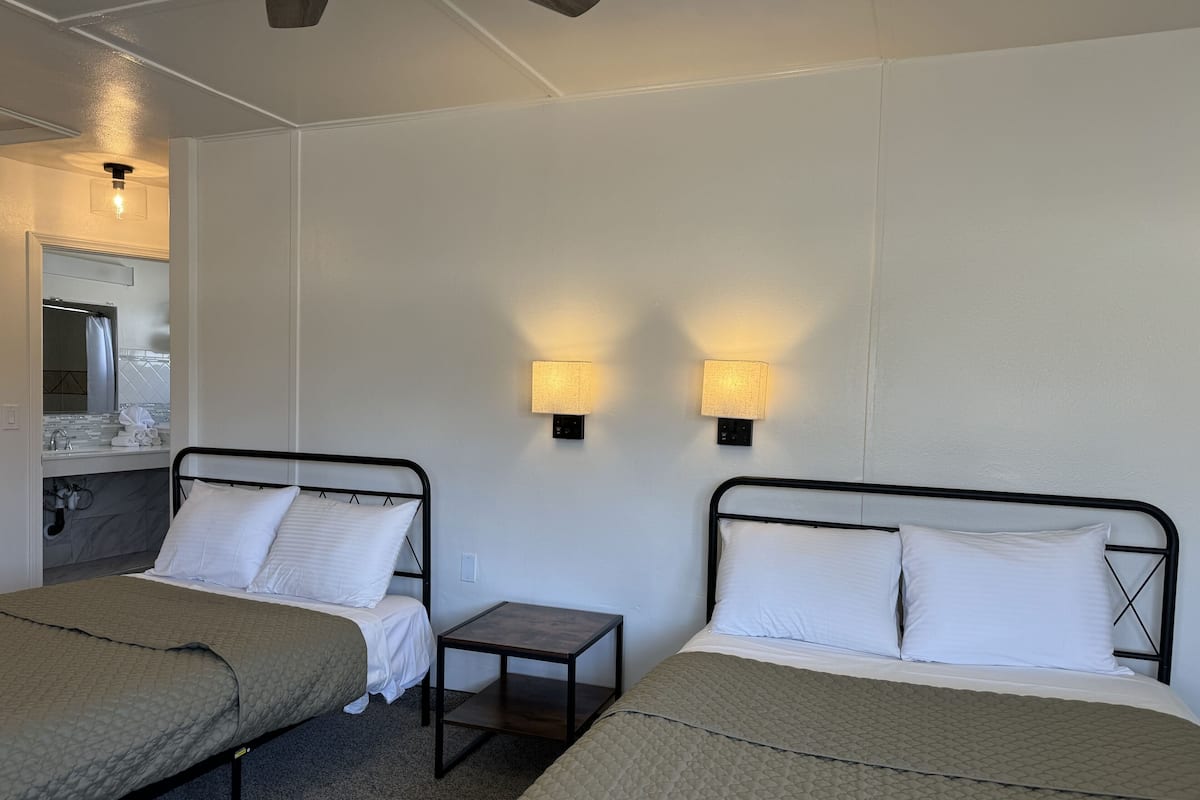 A hotel room with two single beds, a small bedside table, a nightstand, a wall-mounted lamp, and a bathroom with a mirror and towel rack.