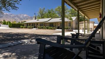 A motel with a covered outdoor seating area, a parking lot, and a mountain backdrop.
