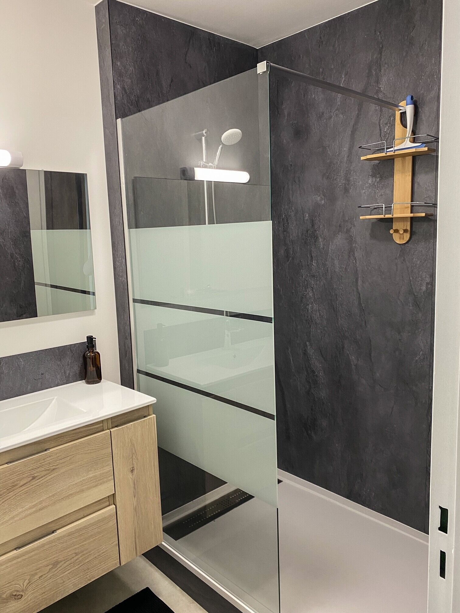 Un baño moderno con cabina de ducha de vidrio, estante de madera con artículos de aseo y un espejo grande sobre el lavabo.