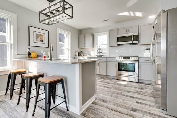 A modern kitchen with a central island, bar stools, stainless steel appliances, and a large window providing natural light.