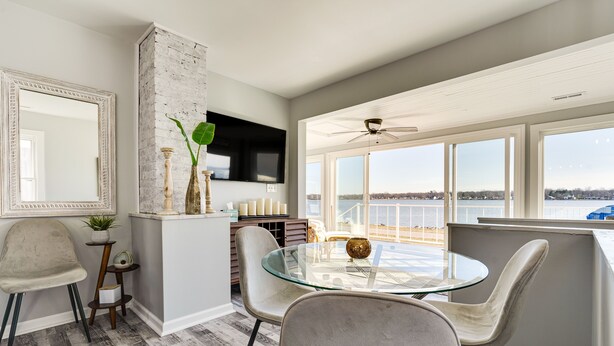 A dining area with a glass table and chairs, a television mounted on the wall, and a view of the water outside.