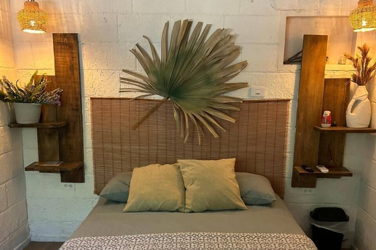 Ein Schlafzimmer mit einem gemusterten Bettüberwurf, einer großen Kübelpflanze an der Wand und Holzregalen mit Dekoration.